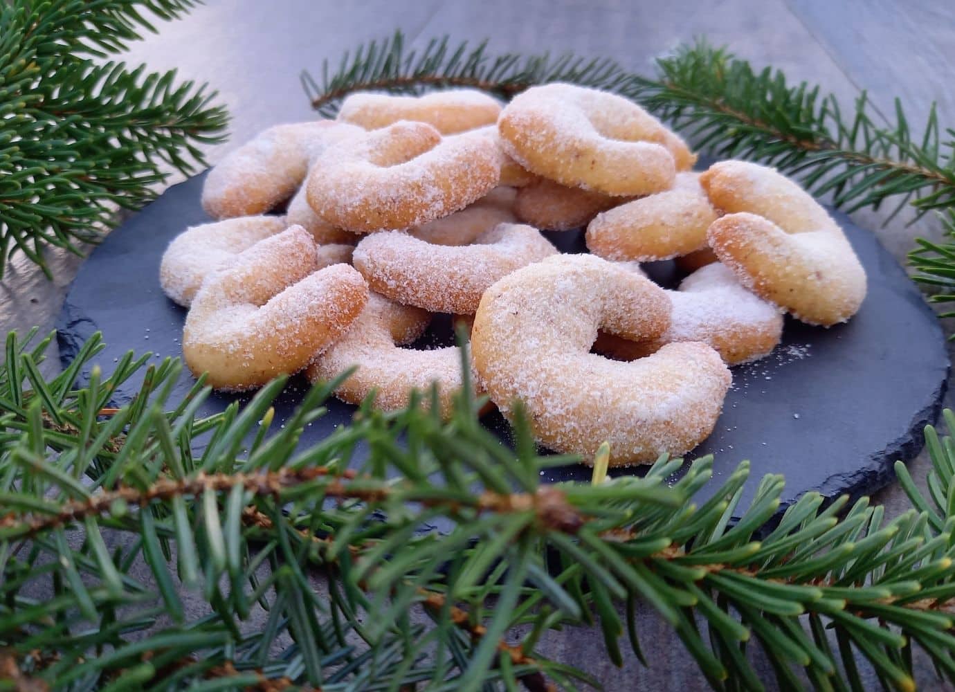Zuckerbestäubte Kekse auf Schieferplatte mit Tannenzweigen.