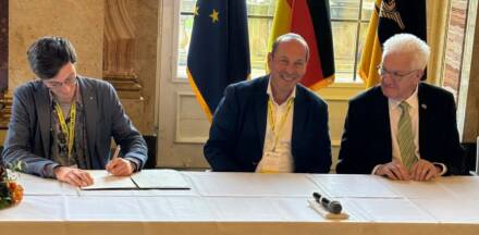 Drei Männer bei einer Vertragsunterzeichnung mit Flaggen.